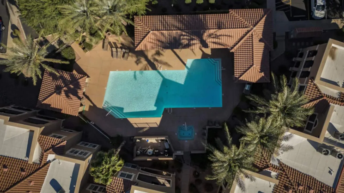 This image captures the community's inviting swimming pool, framed by lush palm trees and adjacent patios, with glimpses of surrounding building rooftops. The scene is bathed in warm, golden light, suggesting a leisurely afternoon or evening ideal for socializing or unwinding.