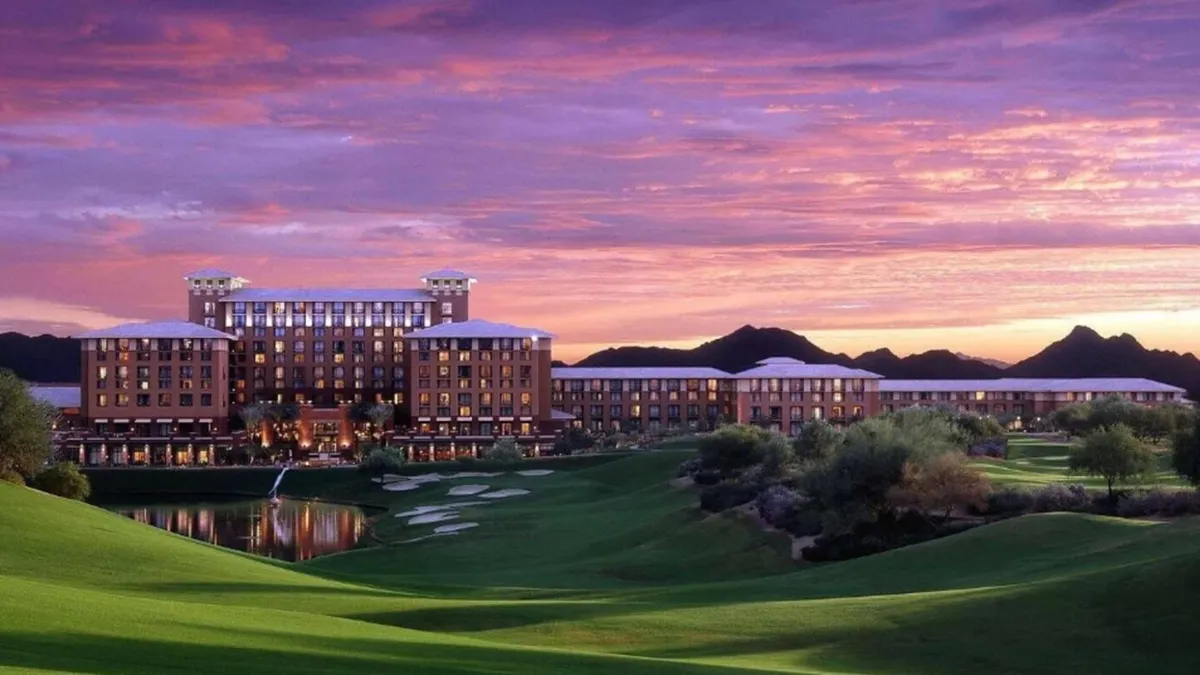 An elegant exterior shot of a resort-like building overlooking a stunning golf course, with a tranquil water feature and a glowing sunset casting warm hues across the sky. The ambiance is both upscale and relaxed, suggesting the sophisticated yet laid-back lifestyle the area offers.