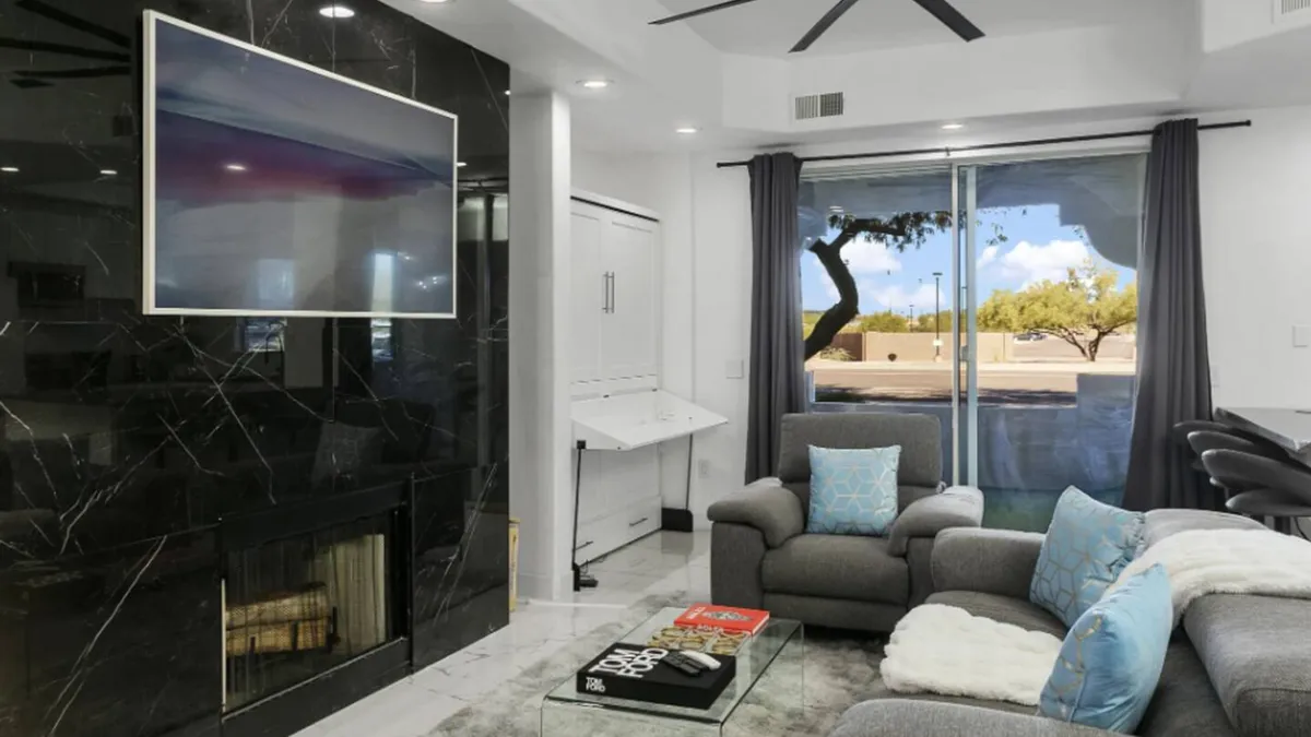 A stylish living room featuring a large television, sleek fireplace, comfortable seating, and a glass coffee table. Sunlight streams in through the window, brightening the space and emphasizing a cozy, contemporary mood ideal for relaxing or socializing.