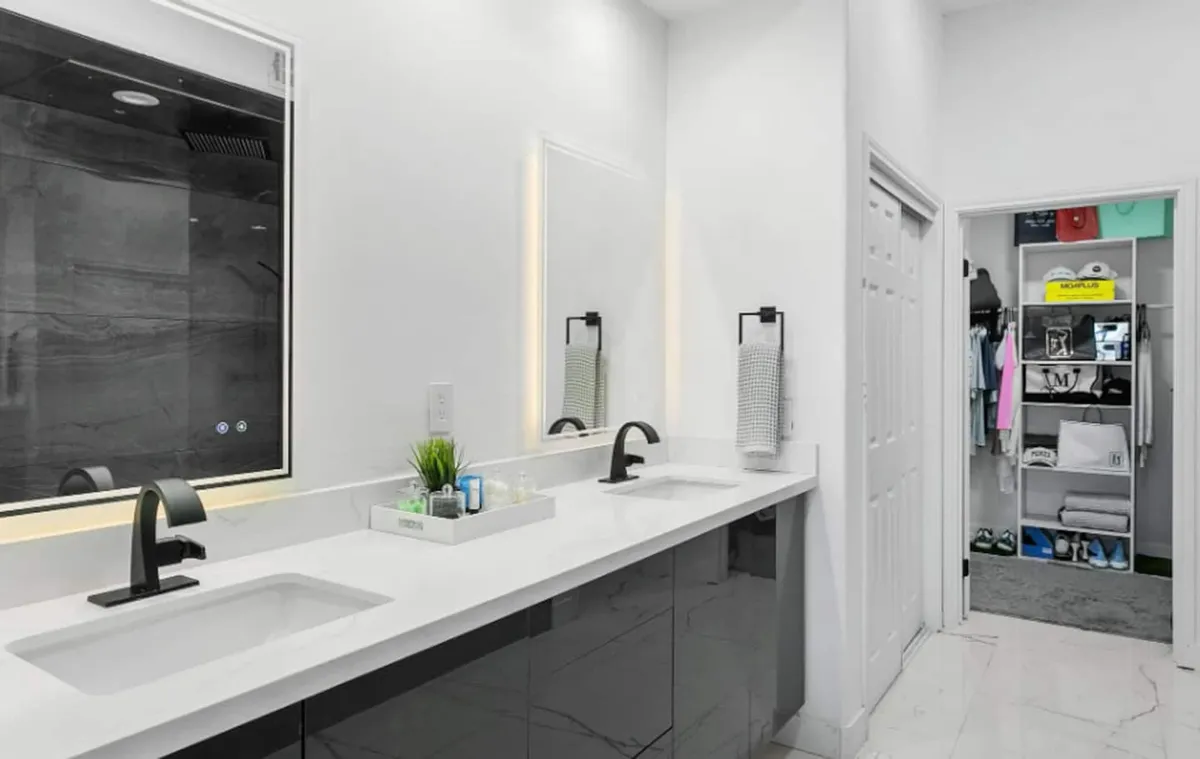 A bright, modern bathroom featuring dual sinks, a spacious countertop, a large mirror, and an organized closet. Sunlight streams in, creating a fresh and inviting atmosphere ideal for personal grooming and relaxation.