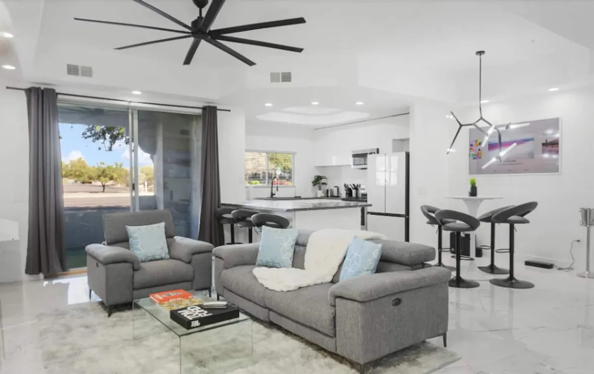 The hero image showcases a stylish and inviting open-concept living space. A plush gray sofa faces a sleek glass coffee table, with a modern kitchen island just beyond, perfect for both relaxation and social gatherings. Soft lighting and tasteful decor create a warm, welcoming atmosphere ideal for unwinding after a day exploring Scottsdale.