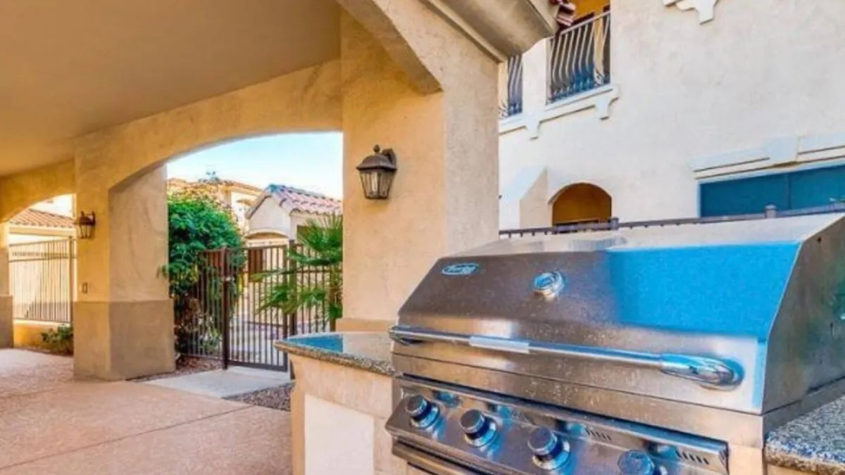 This exterior image features a welcoming patio area with a stainless steel grill, an arched entryway, lush green foliage, and a decorative lantern. The setting is ideal for barbecuing, outdoor dining, and relaxed social gatherings, with an inviting, resort-like ambiance.