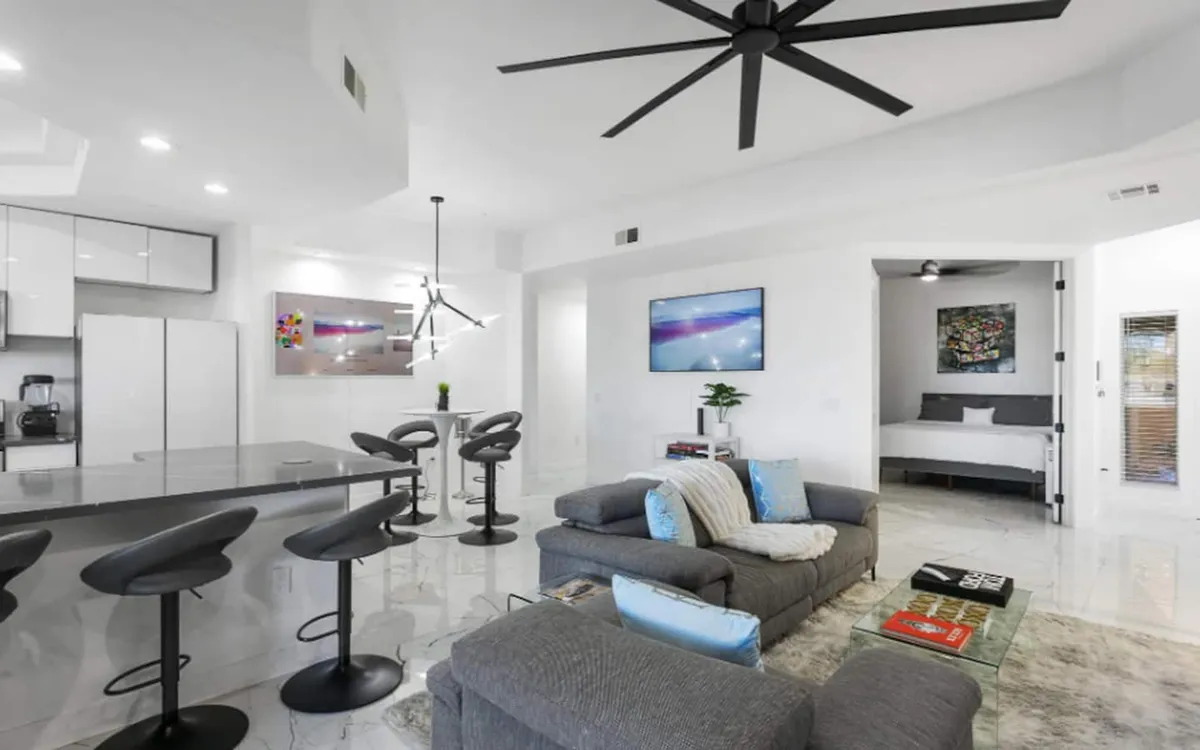 Open-plan living and kitchen area with a kitchen island, modern bar stools, a comfortable sofa with plush cushions, artwork on the walls, and a television. The design encourages social gatherings, casual dining, and relaxation.
