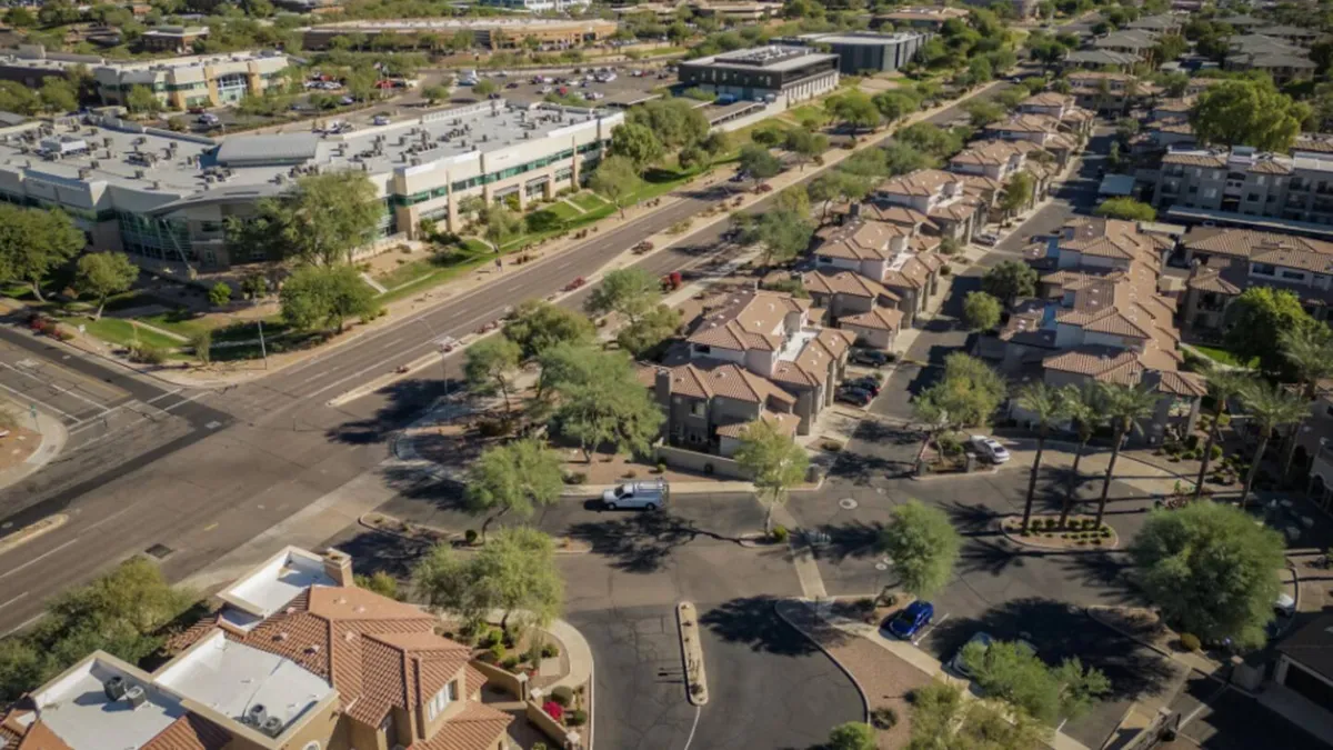 This image features residential and commercial buildings along tree-lined streets, with people walking, vehicles on the road, and a relaxed, community-oriented atmosphere. The mix of buildings and visible activity suggests a lively neighborhood setting convenient for both work and leisure.