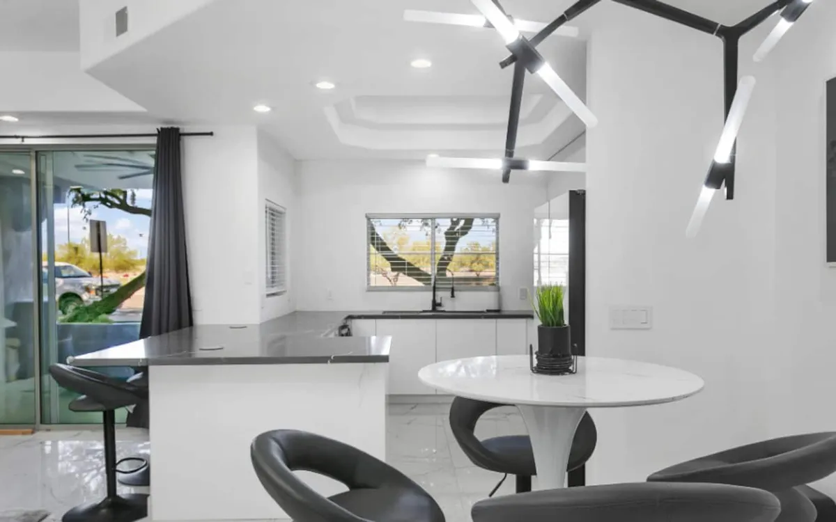 A contemporary dining area with a round table, black bar stools, a striking modern chandelier, and a large window offering an outdoor view. The scene is set for casual dining or entertaining guests, with natural light enhancing the stylish ambiance.