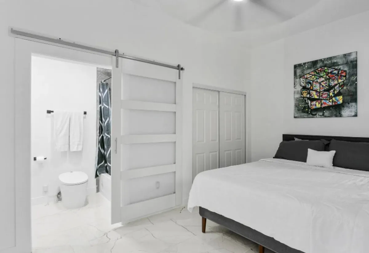 A serene bedroom with a comfortable bed, inviting linens, tasteful artwork, and a visible entrance to the adjoining bathroom. The soft lighting and organized space convey tranquility and personal comfort while providing a glimpse of the ensuite convenience.