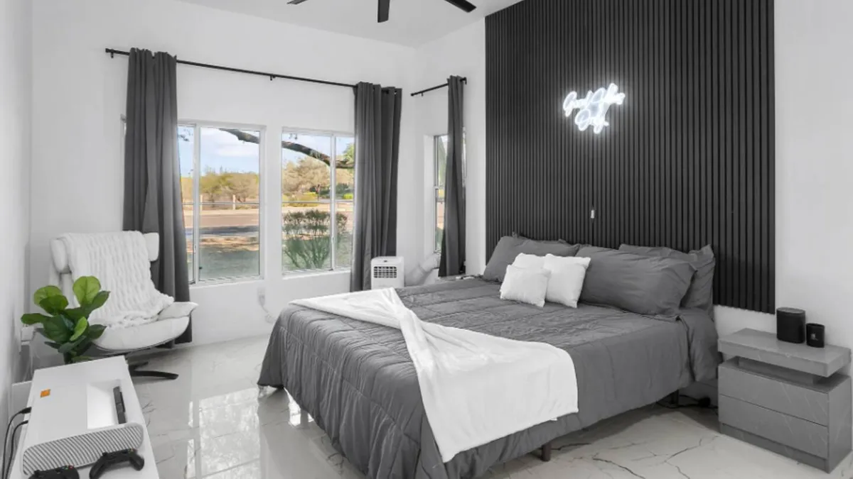 This image showcases a stylish bedroom featuring a bed with gray bedding, a modern neon light sign above the bed, expansive windows providing natural light and views, and a contemporary chair positioned for reading or relaxing. The setting is uncluttered but thoughtfully decorated, giving a sense of calm and comfort.